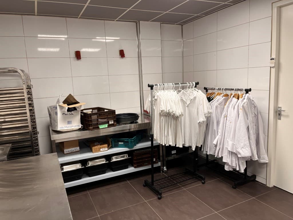 Keukenruimte met stalen werkblad, rekken met witte kleding en diverse opslagmanden aan de wand.