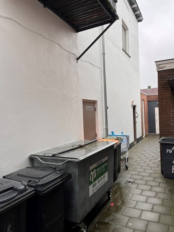Achterzijde van het appartement met vuilcontainers en een betegeld pad, muren zijn wit geschilderd, overdekte afvoer zichtbaa