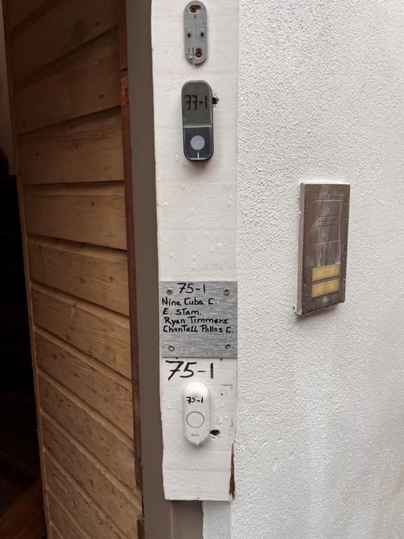 De gevel van een appartement met huisnummers en intercom in een houten omgeving
