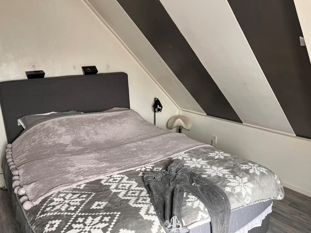 Slaapkamer met schuin plafond, twee nachtlampen en bed met dekbed en plaid op de vloer.