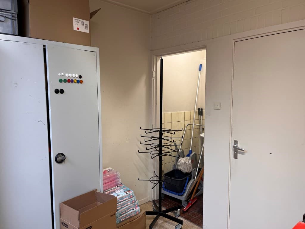In een appartement is een ruimte met een kast, dozen, en een kledingrek bij een doorgang naar de schoonmaakruimte zichtbaar