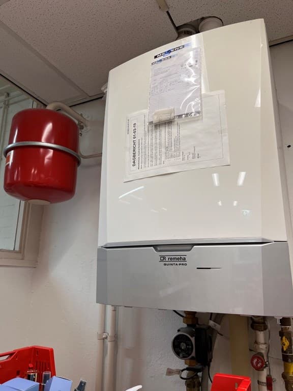 Huiselijke ruimte met een witte ketel, een rode expansietank en verschillende leidingen zichtbaar aan de muur.