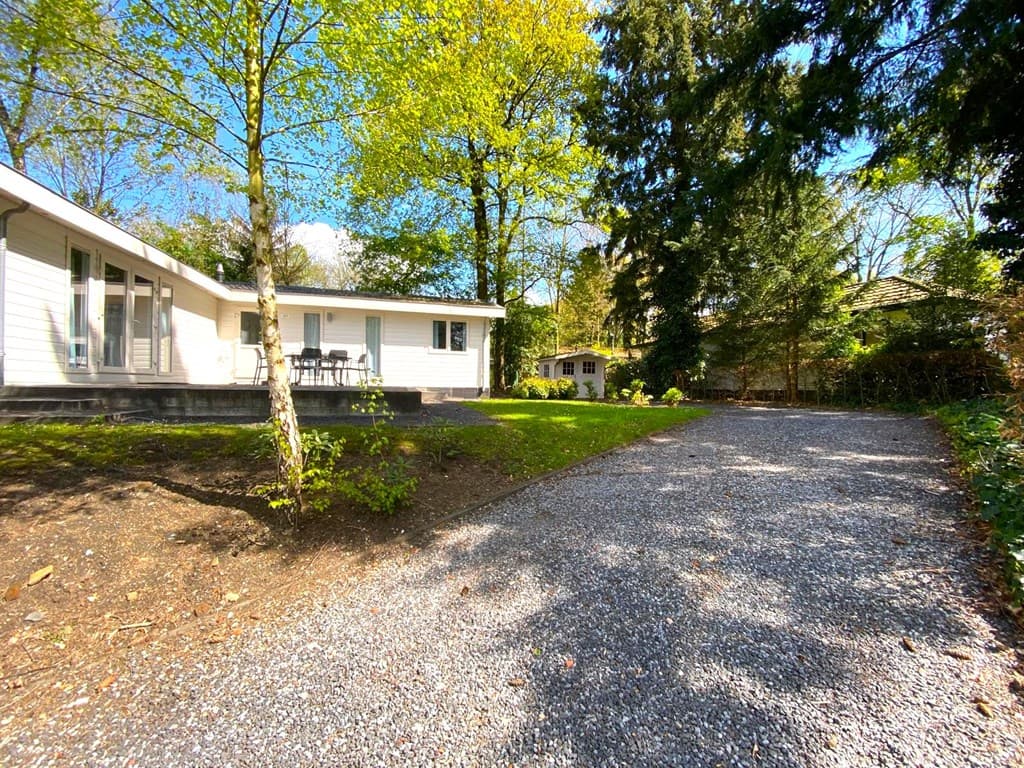 Moderne vakantiewoning omgeven door groen, met een grindpad en terrasgedeelte aan de zijkant.
