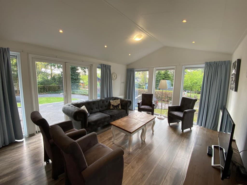 Lichte woonkamer met een bank, fauteuils, salontafel en grote ramen met uitzicht op groen buiten.