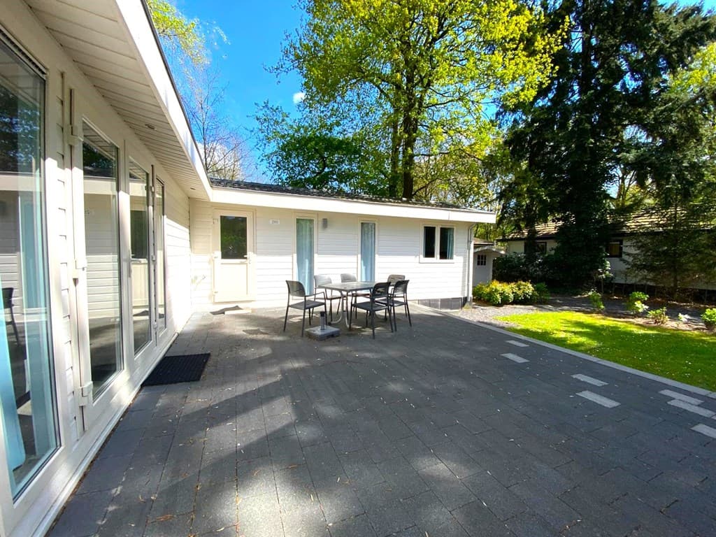 Moderne vakantiewoning met een betegeld terras en een tafel met stoelen, omringd door groene bomen.