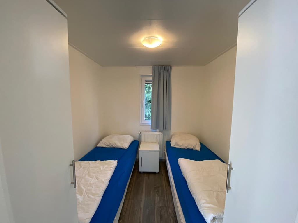 Slaapkamer met twee eenpersoonsbedden, witte muren en een raam met een grijze gordijn.