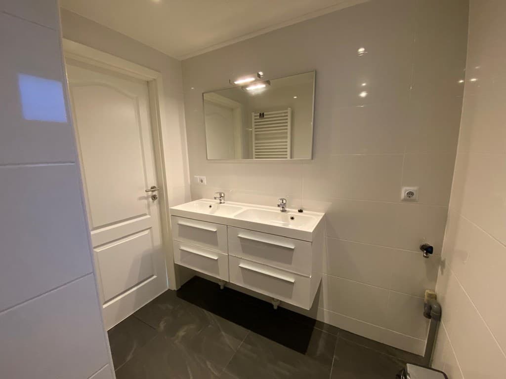 Lichte badkamer met dubbele wastafel en spiegel, witte betegeling en donkere vloer.