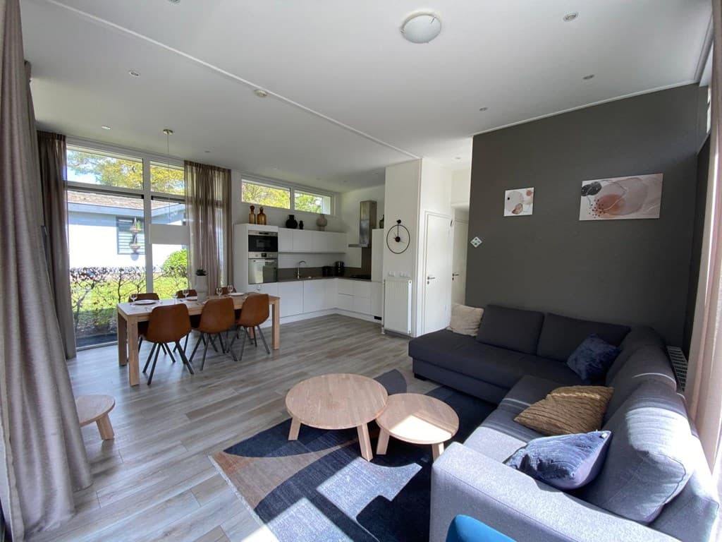 Moderne woonkamer met open keuken, eethoek en zithoek met banken en tafel, veel natuurlijk licht vanuit de ramen.