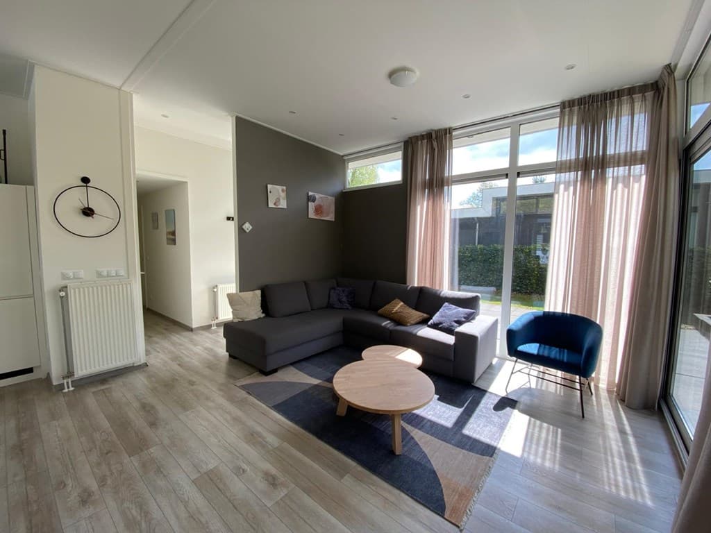 Moderne woonkamer met grote ramen, een grijze bank, een houten salontafel en een blauwe stoel.