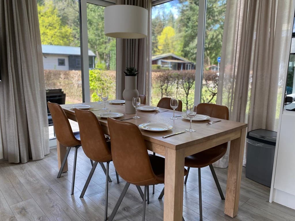 Eettafel met zes stoelen, grote ramen met uitzicht op groen, lichte inrichting en een moderne hanglamp.