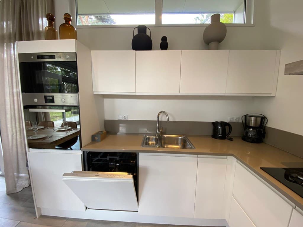 Moderne keuken met witte kasten, rvs spoelbak en verschillende apparatuur, zoals een oven en vaatwasser.