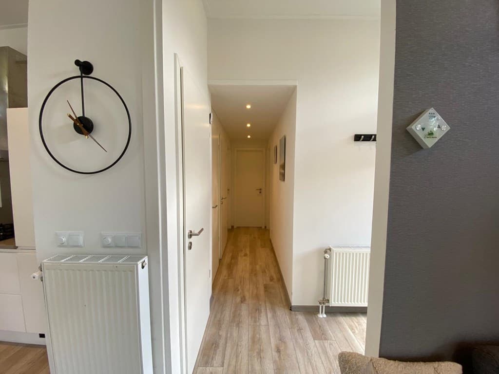 Aanzicht van een smalle gang met een klok aan de muur, radiatoren en een deur aan het einde.