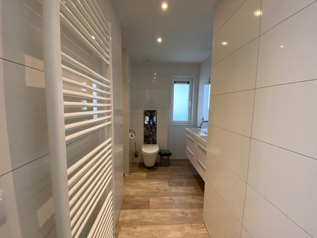 Moderne badkamer met witte tegels, inloopdouche, toilet en wastafel met kast. Verwarmde handdoekradiator zichtbaar.