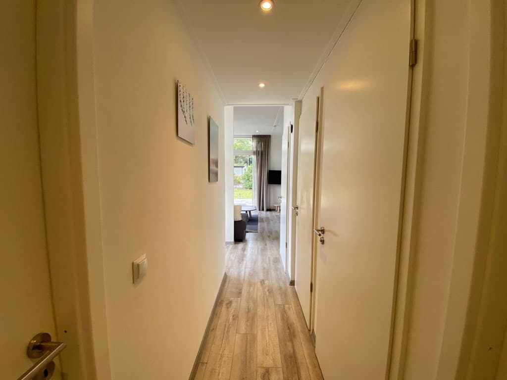 Dichte gang met lichte muren en een houten vloer, leidt naar een woonkamer met een bank en een tv.