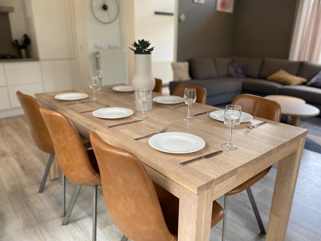 Eetkamertafel met zes stoelen, gedekt met borden en glazen, naast een woonkamer met een bank en een plantenpot