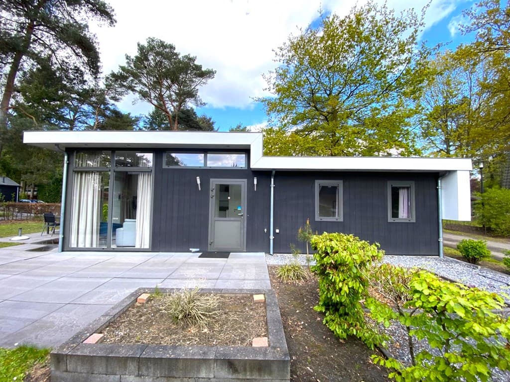 Moderne vakantiewoning met houten gevel en glazen wand, grote terrastegels en groene beplanting rondom.