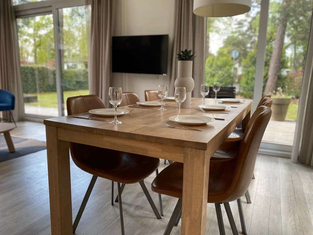 Eettafel met houten blad en bruine stoelen, glazen en borden klaar voor een maaltijd, uitzicht op de tuin achter het huis.
