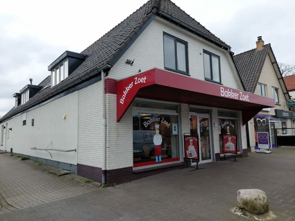 Hoekpand met een witgekalkte gevel en een rode luifel, met grote ramen en een ingang aan de voorkant.