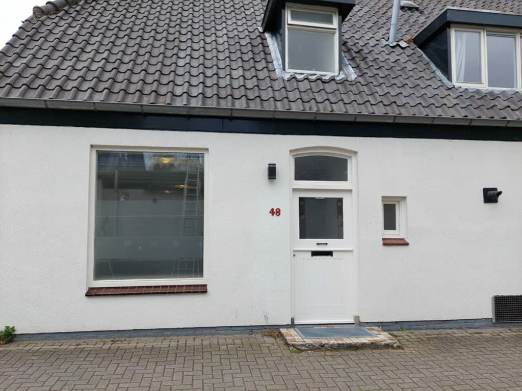 Witte gevel met grijze dakpannen en een voordeur met bovenlicht, naast grote ramen met ventilatieroosters.