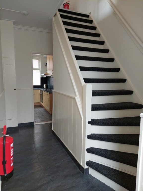 Trap met zwarte bekleding en witte leuning, hal met vloer in grijstinten en deur naar keuken aan het eind van de gang