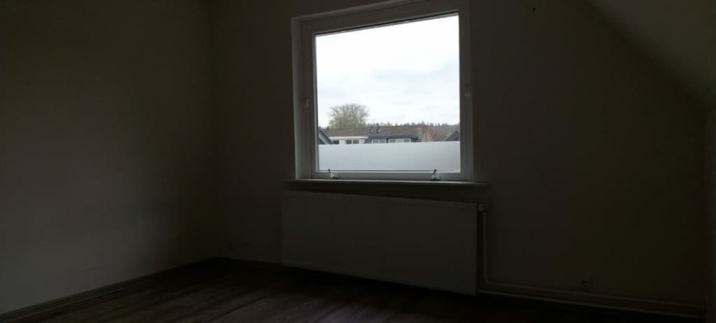 Ruime kamer met een groot raam en een verwarmingsradiator, lichte muren en een laminaatvloer.