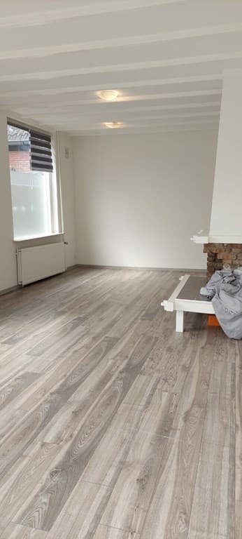 Ruime woonkamer met lichte houten vloer, witte muren en een open haard in de hoek.