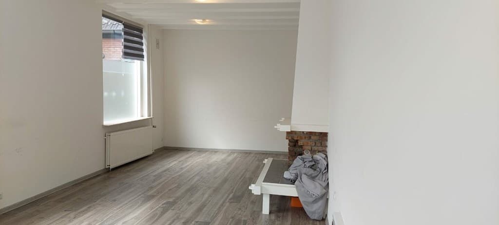 Lichte woonkamer met houten vloer, witte muren en een schoorsteen aan de rechterkant, zonder meubels.