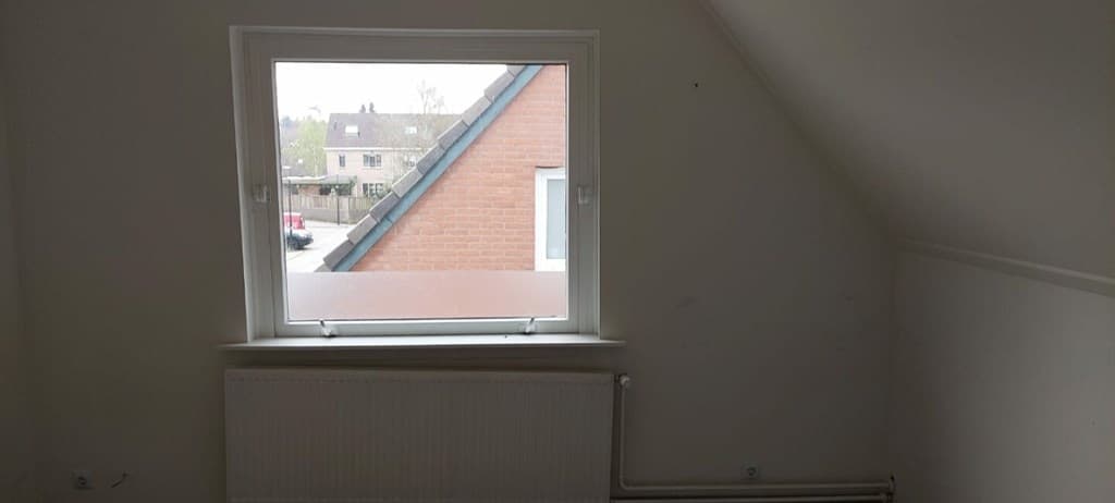 Zicht op een raam met uitzicht op een straat en een huis met een bakstenen gevel, witte muren en verwarmingselement zichtbaar