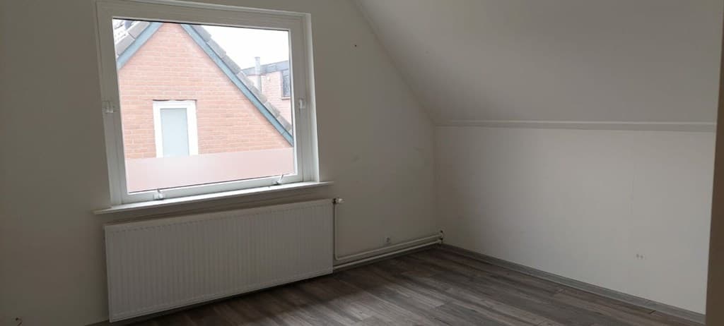 Lege kamer met schuin plafond, grote raam met uitzicht op aangrenzende gevel en houten vloer.