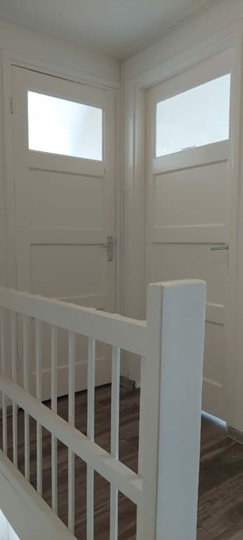 Binnenaanzicht van een gang met twee gesloten deuren en een witte leuning. Houten vloer en lichtinval door de ruiten.