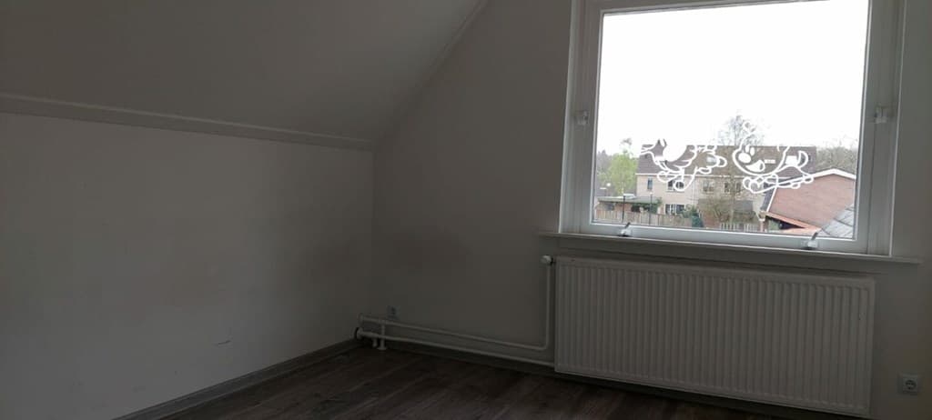 Lege ruimte met schuin dak en een raam dat uitzicht biedt op groen en omliggende woningen.