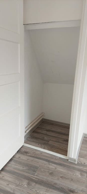 Kleine ruimte met schuine wanden en houten vloer, met een deur en zichtbaar leidingwerk aan de muur