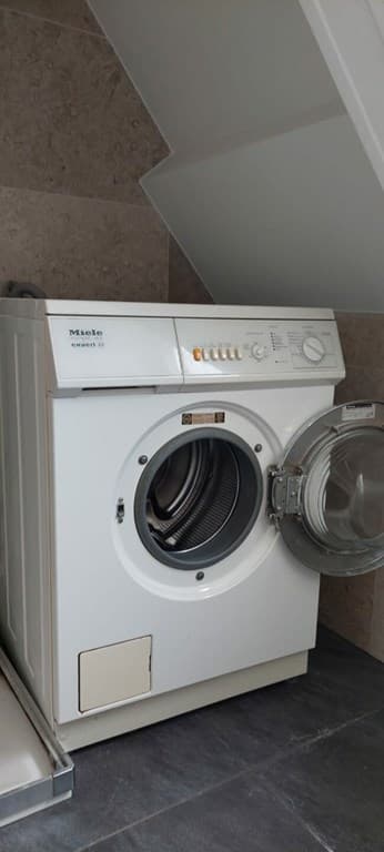 Wit wasmachine van Miele in een hoek met een tegelvloer en een schuine wand.