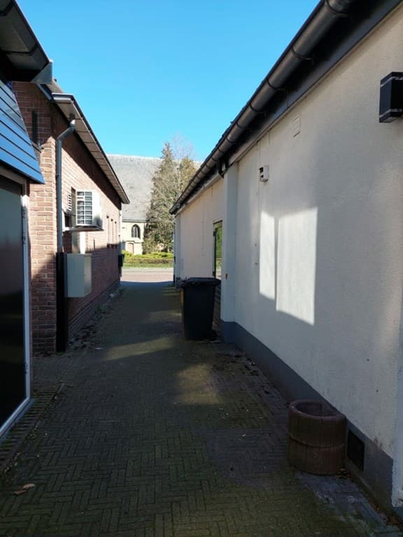 Smalle doorgang tussen twee huizen met een vuilnisbak en een helder blauwe lucht boven.