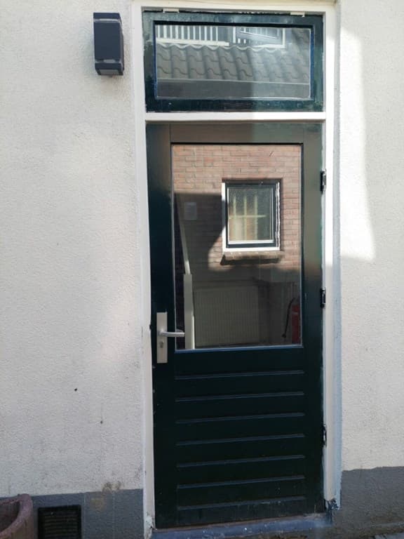 Zwarte houten deur met bovenraam, bevestigd aan een lichte muur met een klein venster aan de zijkant.