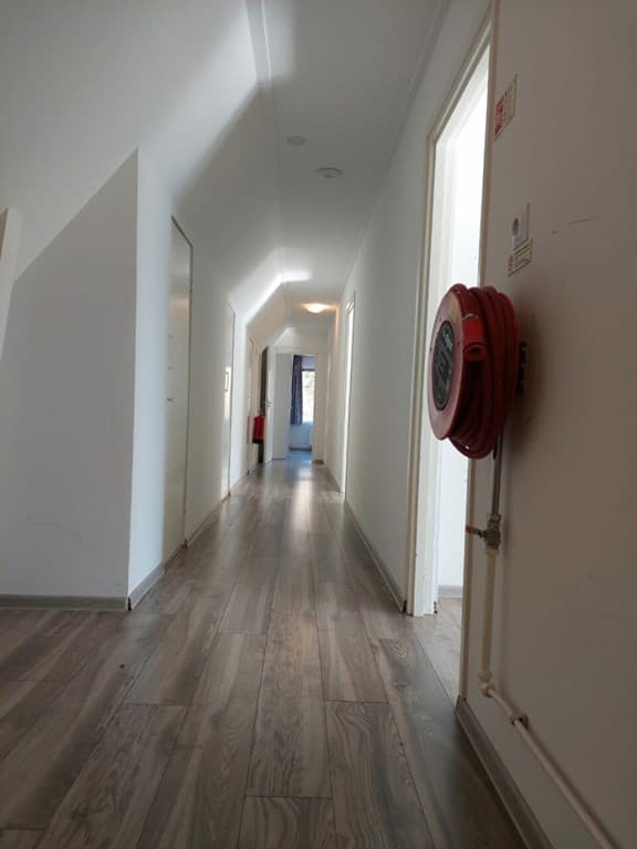 Langgerekte gang met houten vloer, witte muren en deuren aan beide zijden van de ruimte, met een brandblusser aan de muur.