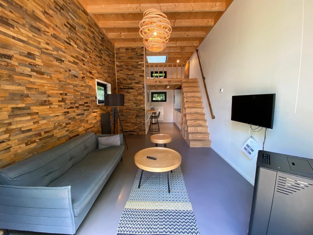 Hedendaagse woonkamer met houten wand, bank, salontafels en open ruimte naar de keuken en trap naar boven.