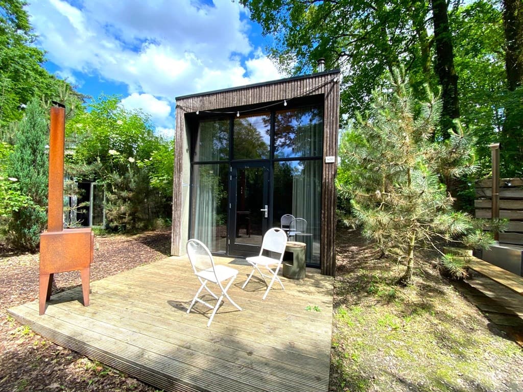 Moderne vakantiewoning met grote ramen, houten terras en twee stoelen in een groene omgeving.