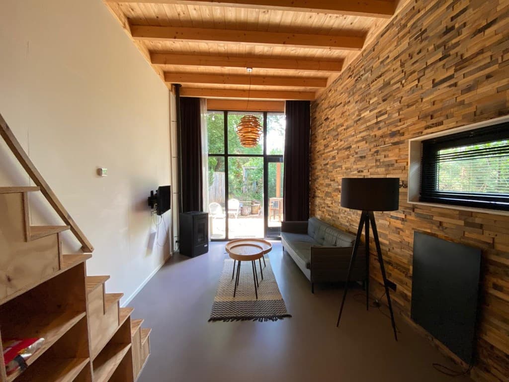 Ruime woonkamer met houten plafonds, een houten wand, bank, lamp, tv en uitzicht op het terras en de tuin.