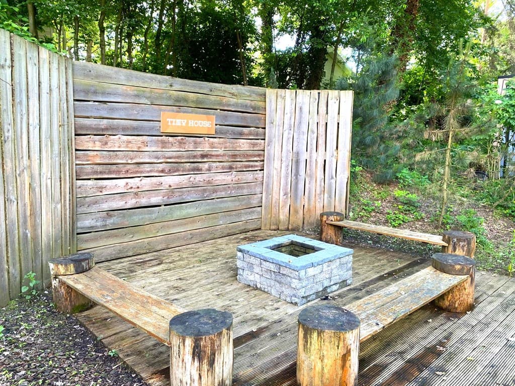 Buitenruimte met houten banken en een vuurplaats, omringd door bomen en een houten schutting.