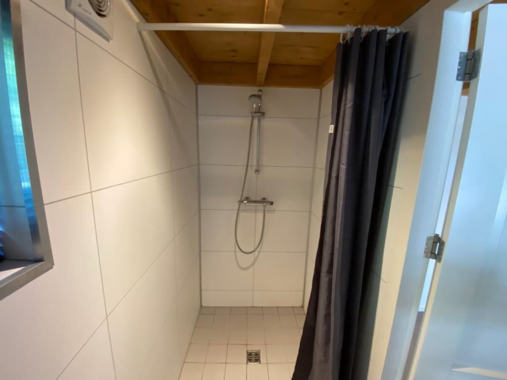 Doucheruimte met grijze tegels, houten plafond en doucherail aan de muur met een gordijn voor privacy.