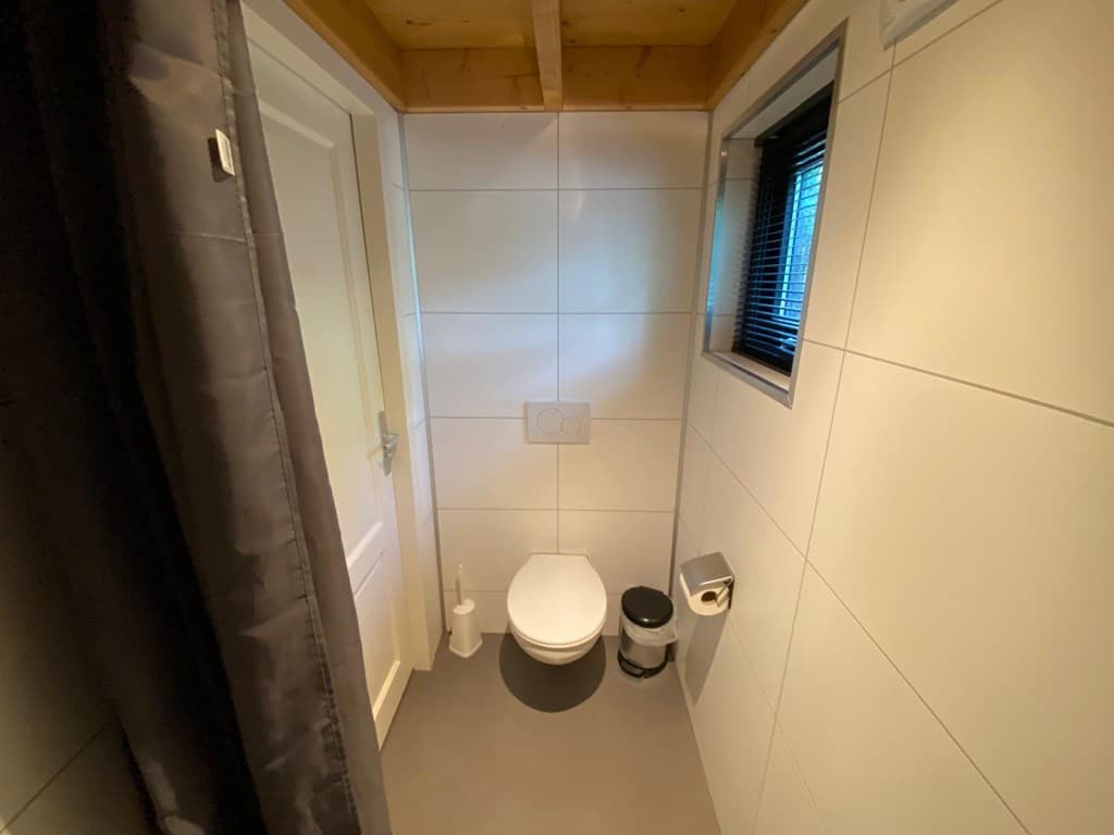 Klein en modern toilet met witte tegels, een toilet en een raam met jaloezieën.