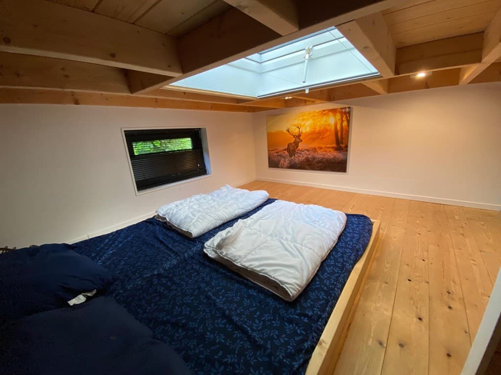 Slaapkamer met houten vloer, hemelraam en kunstwerk van een hert aan de muur. Twee bedden met dekbedden liggen op een verhoog