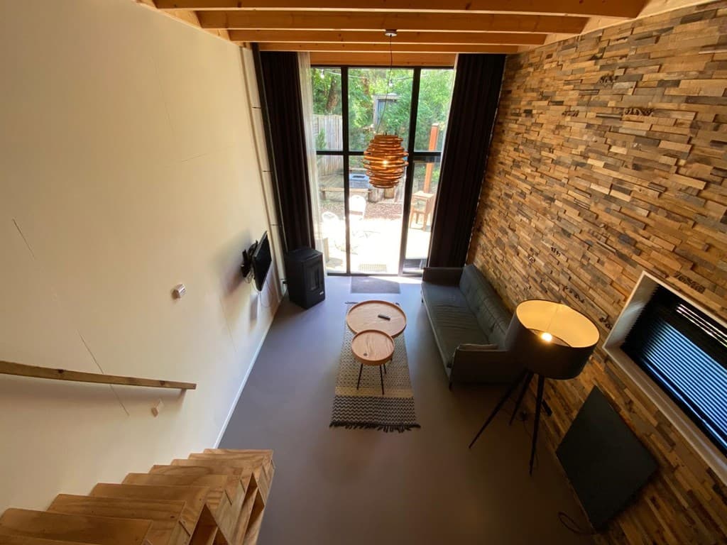 Woonkamer met houten wand, bank, tafels en uitzicht op tuin door grote glazen deuren.