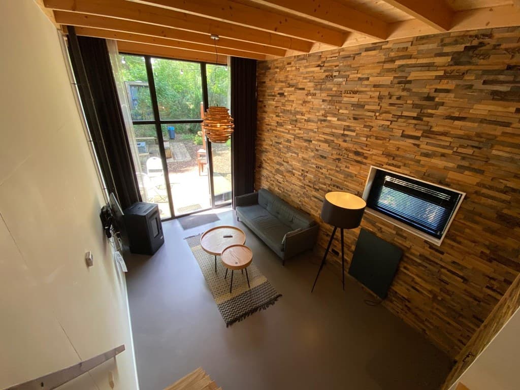 Ingerichte woonkamer met houten muur, bank, lamp en uitzicht op tuin via grote ramen.