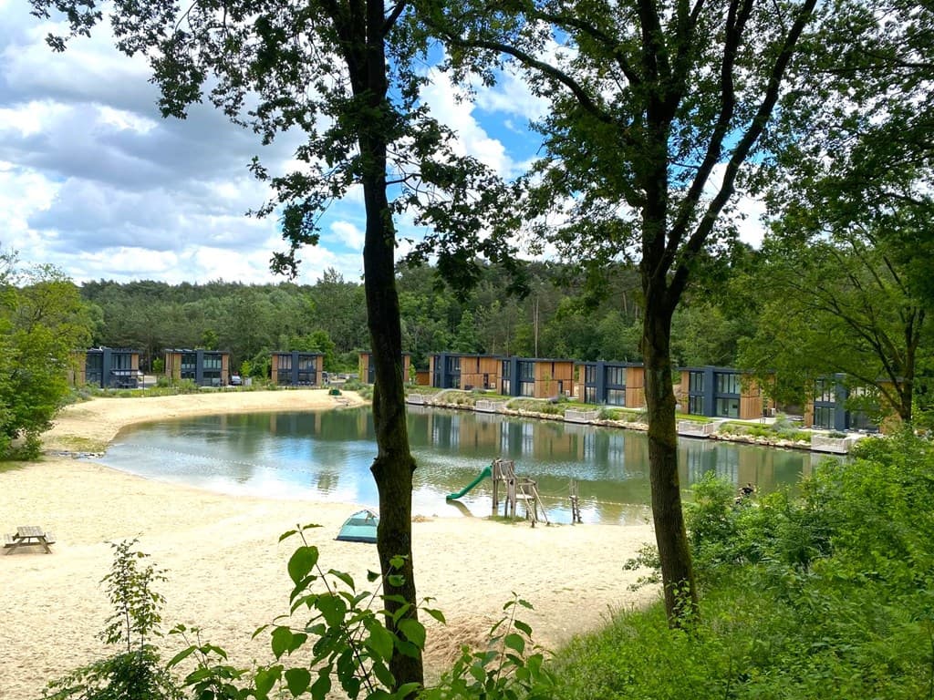 Aanzicht van een waterpartij met zandstrand omringd door bomen en moderne vakantiewoningen in de achtergrond.