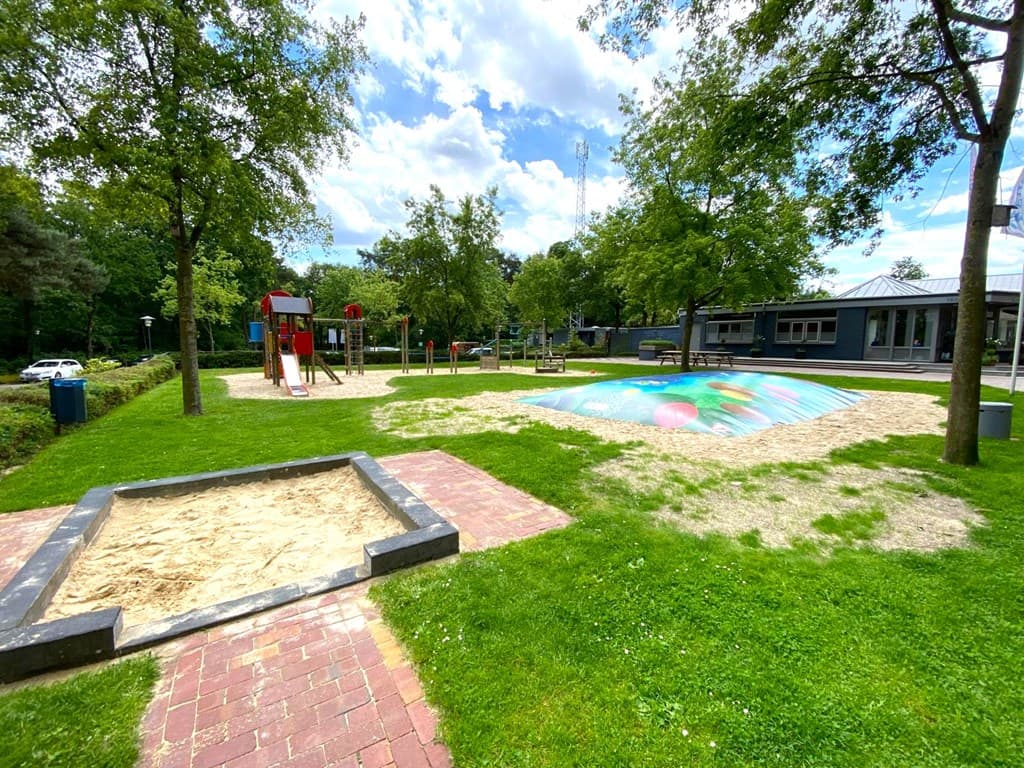 Speeltuin met schommel, glijbaan en zandbak, omringd door gras en bomen, met zwembad op de achtergrond