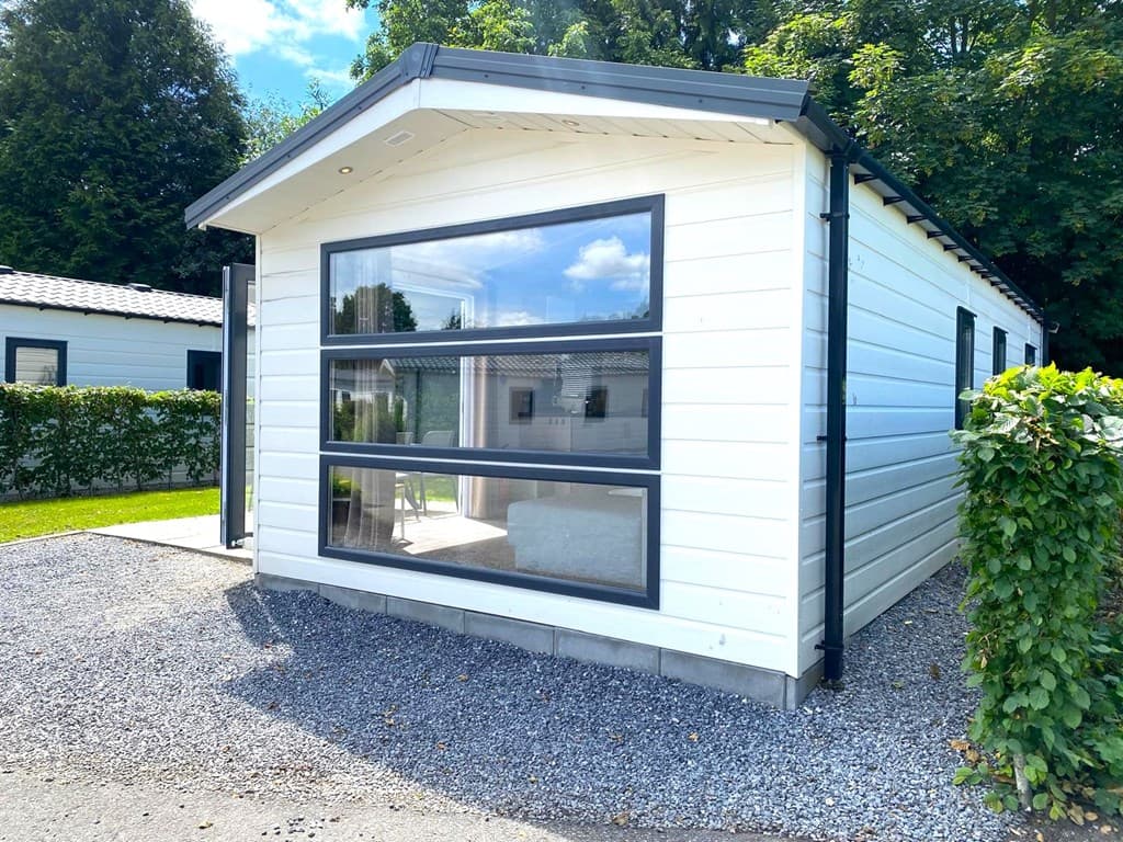 Moderne vakantiewoning met grote ramen en witte gevel, omgeven door groen en grijze kiezelstenen.