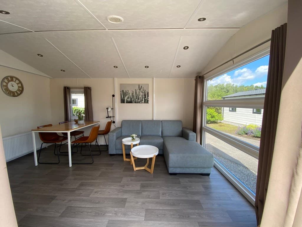 Woonkamer met een grijze bank, een eettafel, en grote ramen met uitzicht op de buitenruimte.