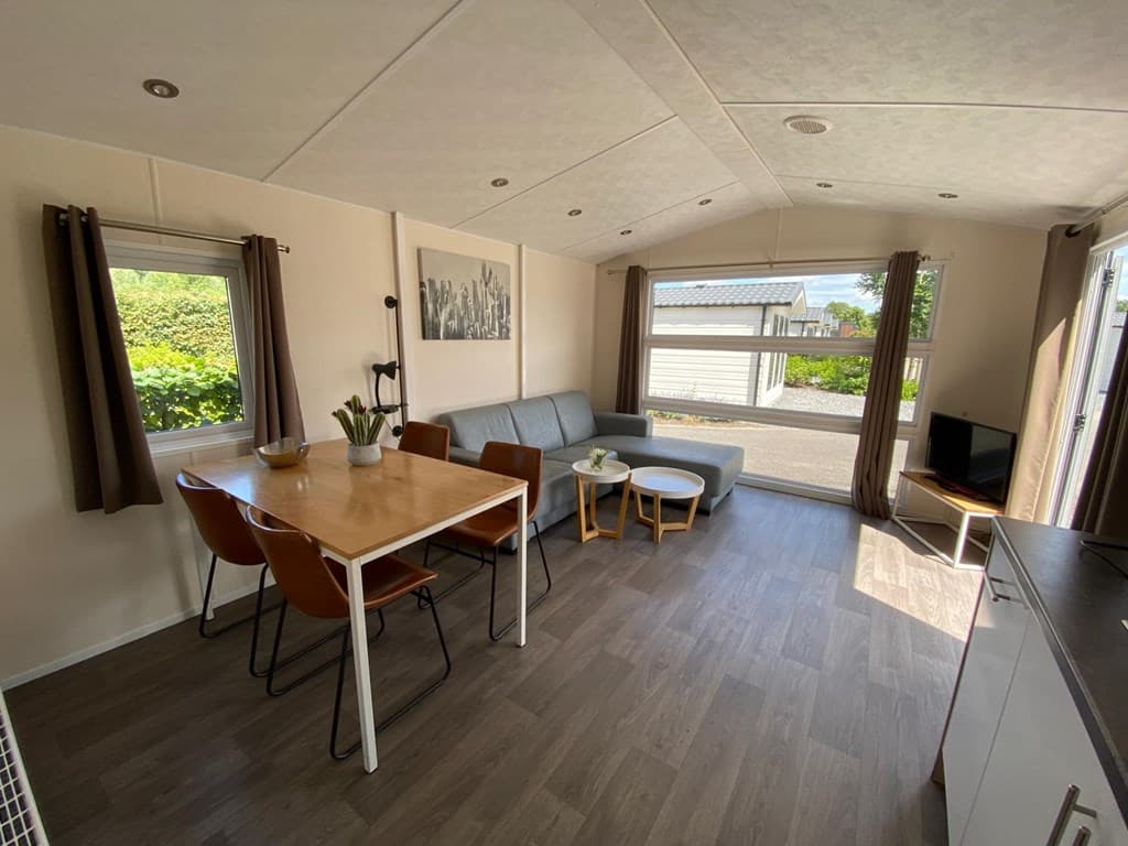 Zithoek met een bank, eettafel met stoelen en openslaande deuren naar buiten in een lichte ruimte.